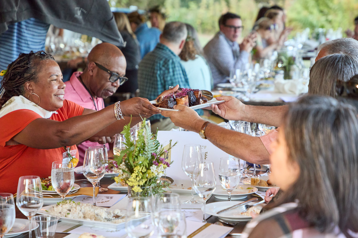 Guests seated at a long table share a plated dish during a lively outdoor farm dinner, surrounded by flowers, glassware, and conversation.
